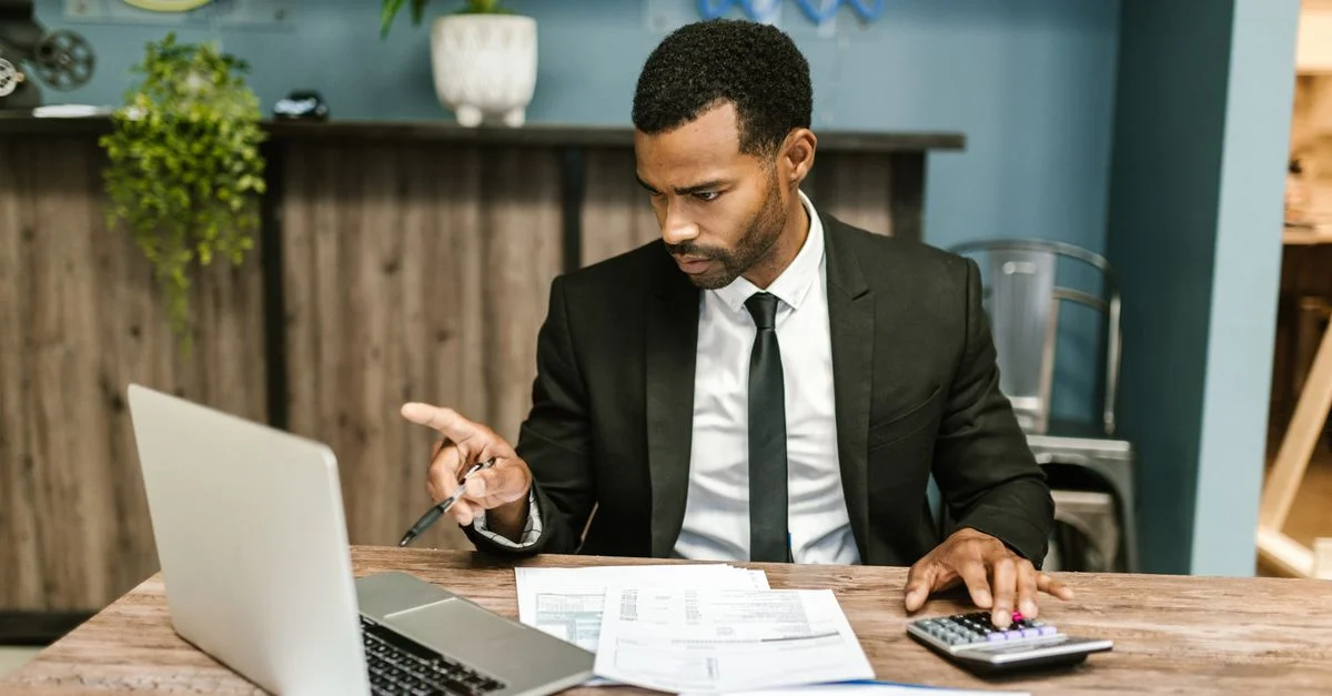 Homme professionnel en costume utilisant un ordinateur portable et une calculatrice dans un cadre de bureau.