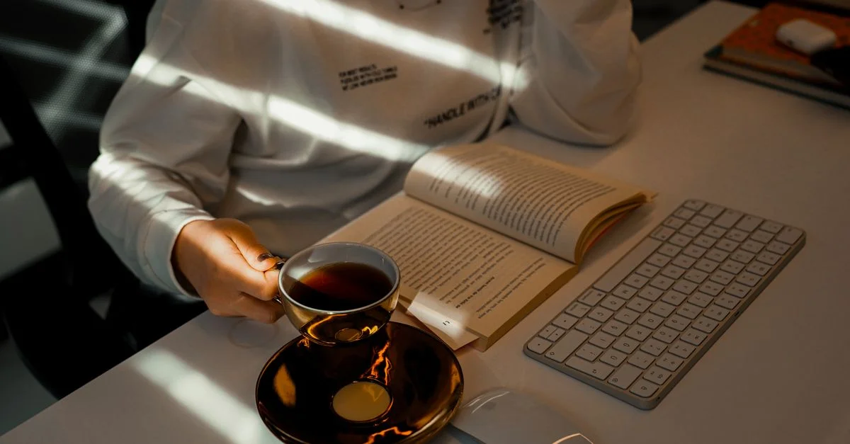 Une femme lisant un livre tout en tenant une tasse de thé à un bureau avec la lumière du soleil qui pénètre.