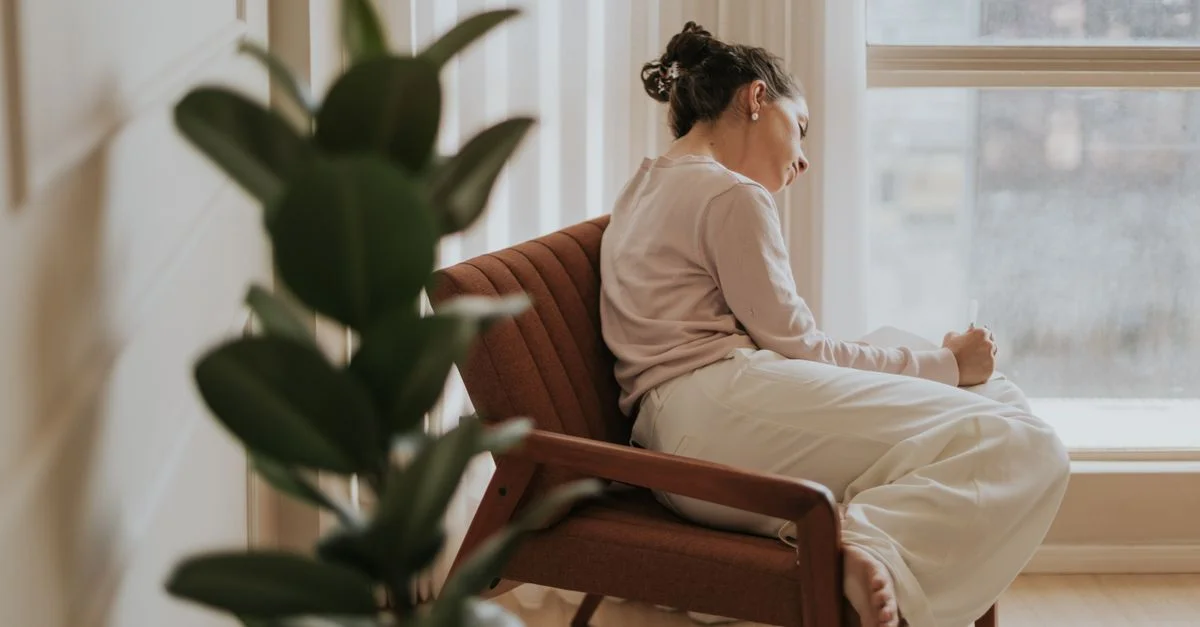 Une femme se détend sur une chaise près d'une fenêtre, plongée dans ses pensées, à l'intérieur.