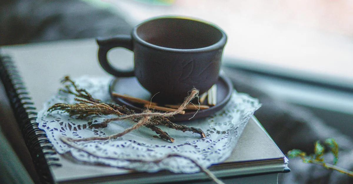 Une tasse en céramique rustique est posée sur des cahiers empilés avec une nappe décorative et des bâtons à proximité.