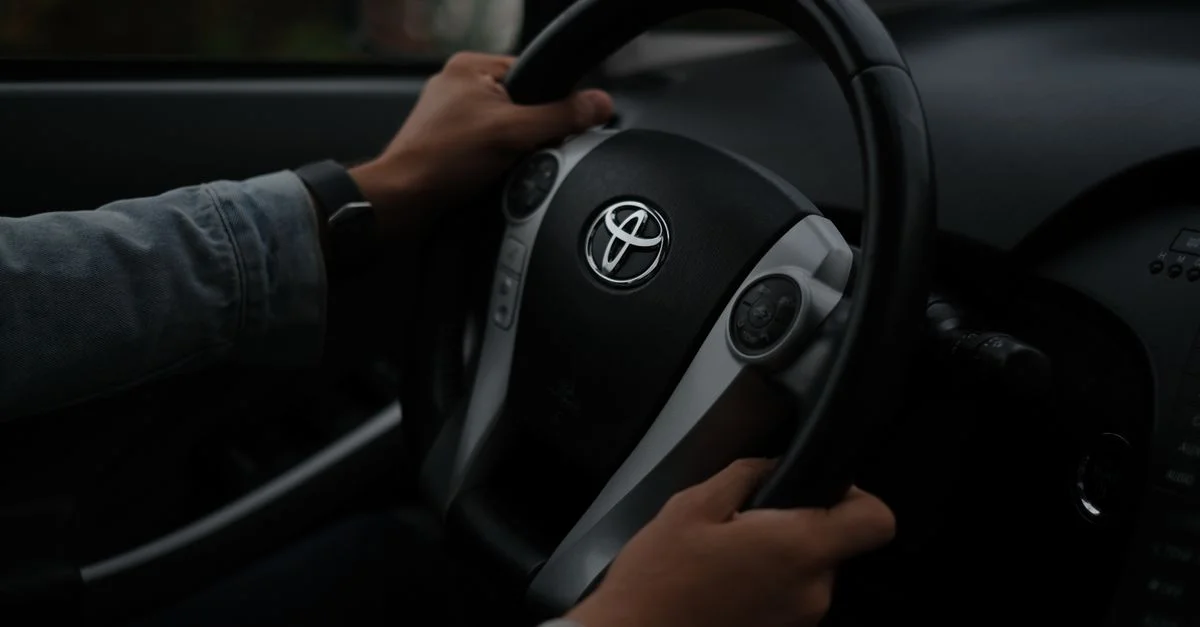 Mains sur un volant Toyota, gros plan à l'intérieur d'un véhicule. Atmosphère sombre.